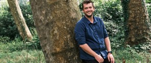 Man in een blauwe blouse leunt tegen een boom in een groen bos.