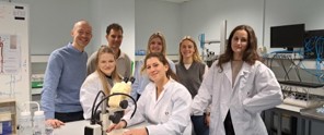 Zes wetenschappers in labjassen staan om een werkplek met een microscoop en glimlachen levendig.