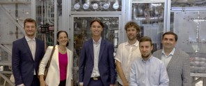 Zes mensen poseren voor een machine in een industriële setting, omringd door apparatuur en technologie.