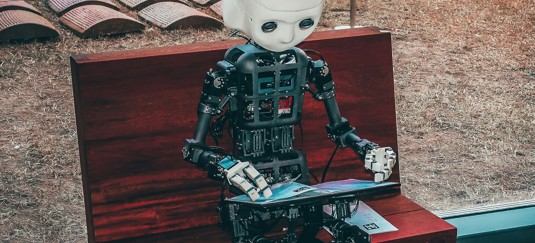 Een robot met een mensachtig gezicht zit op een houten bank en kijkt naar een tablet die hij vasthoudt.