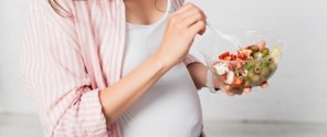 Een zwangere vrouw in een gestreepte blouse houdt een kom salade vast en brengt met een vork een hapje naar haar mond.