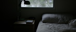 Cozy bedroom with an unmade bed, a nightstand featuring a book and a green lamp, and natural light streaming through the window.