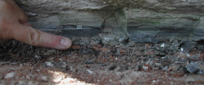 A hand points to a layer of soil and stones beneath a wall, important details for archaeological research.