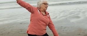 Een persoon in een gestreept rood en wit shirt staat op het strand en wijst naar de zee, met zachte golven op de achtergrond.