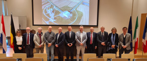 A group of ten individuals in formal attire stands in front of a presentation, with an aerial view of a building and farmland visible in the background.