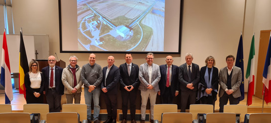 A group of ten individuals in formal attire stands in front of a presentation, with an aerial view of a building and farmland visible in the background.