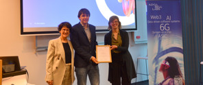 Drie mensen staan op het podium en tonen een certificaat, met een achtergrondbanner die technologie benadrukt.