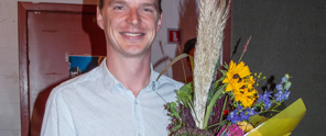 Een persoon in een lichtblauw overhemd houdt een groot boeket met zonnebloemen en andere kleurrijke bloemen vast.