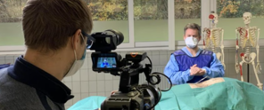 Een man in een operatiekamer legt een medische procedure uit terwijl een cameraman filmt. Op de achtergrond zijn twee anatomische modellen te zien.