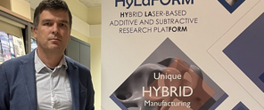 Man staat naast een banner met de tekst 'HyLARFORM Hybrid laser-gebaseerd additief en subtractief onderzoeksplatform' en 'Unieke HYBRIDE Productie'.